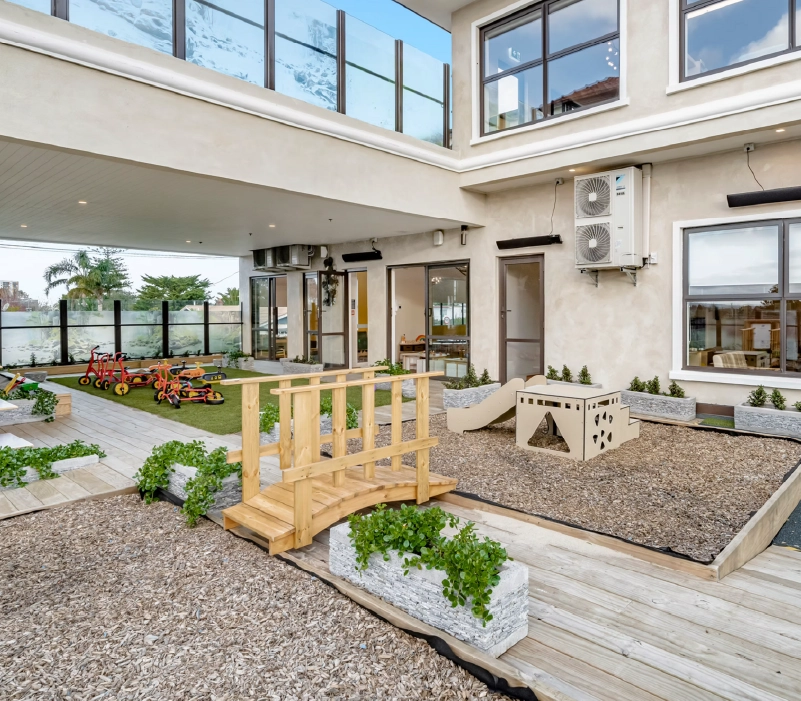 Outdoor play courtyard with timber bridge, modular slide unit, tricycles on turf, and perimeter planters