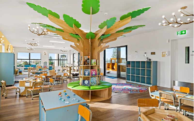 Bright early learning room with tree-form book display, child-height tables and seating, and teal open storage