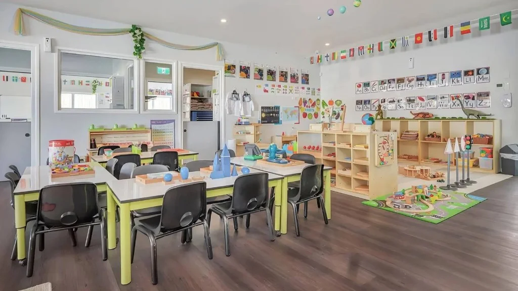 Bright early learning room with child-sized activity tables, small chairs, low wooden shelving, and materials set out for group activities